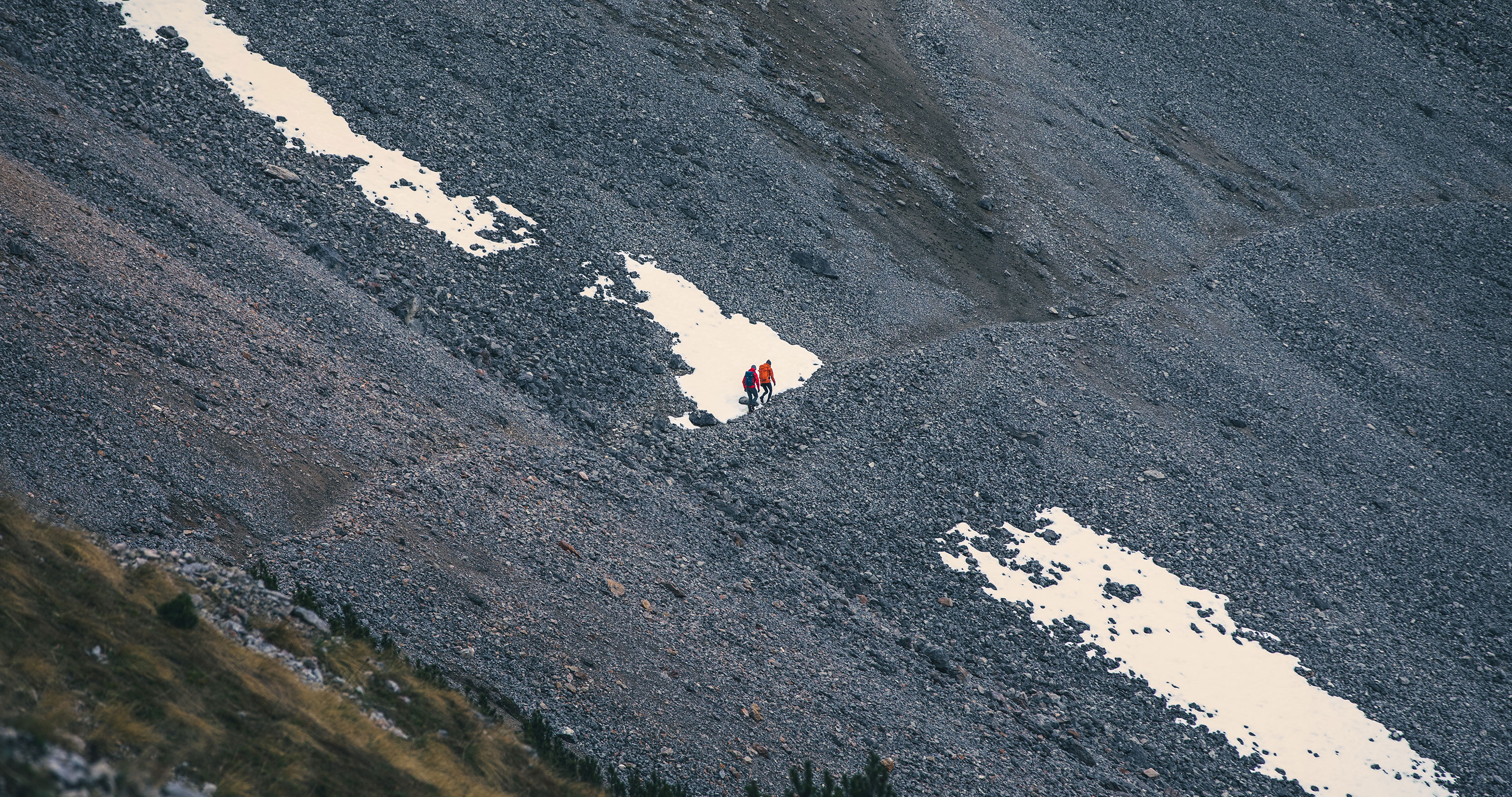 Zwei Wanderer aus der Ferne beim Queren eines Schneefeldes in einem weiten Schotterhang