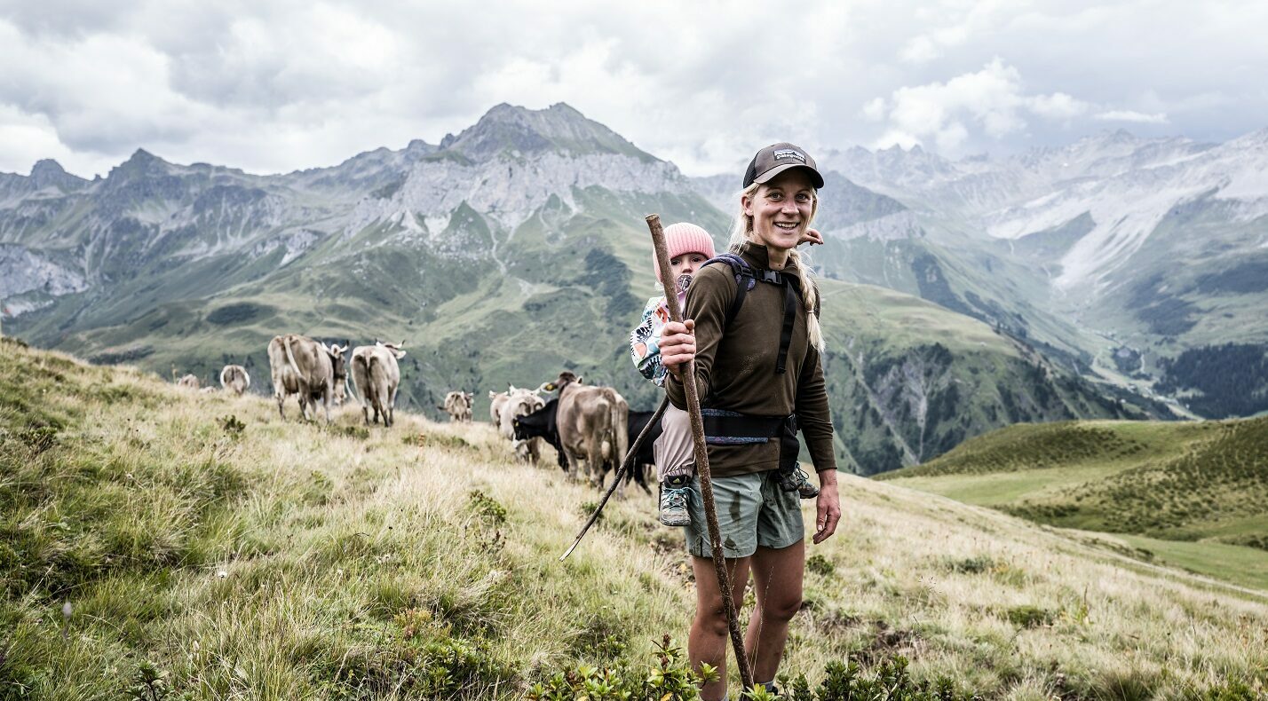Katharina Krepold with her little daughter on her back standing in front of mountains and some cows.