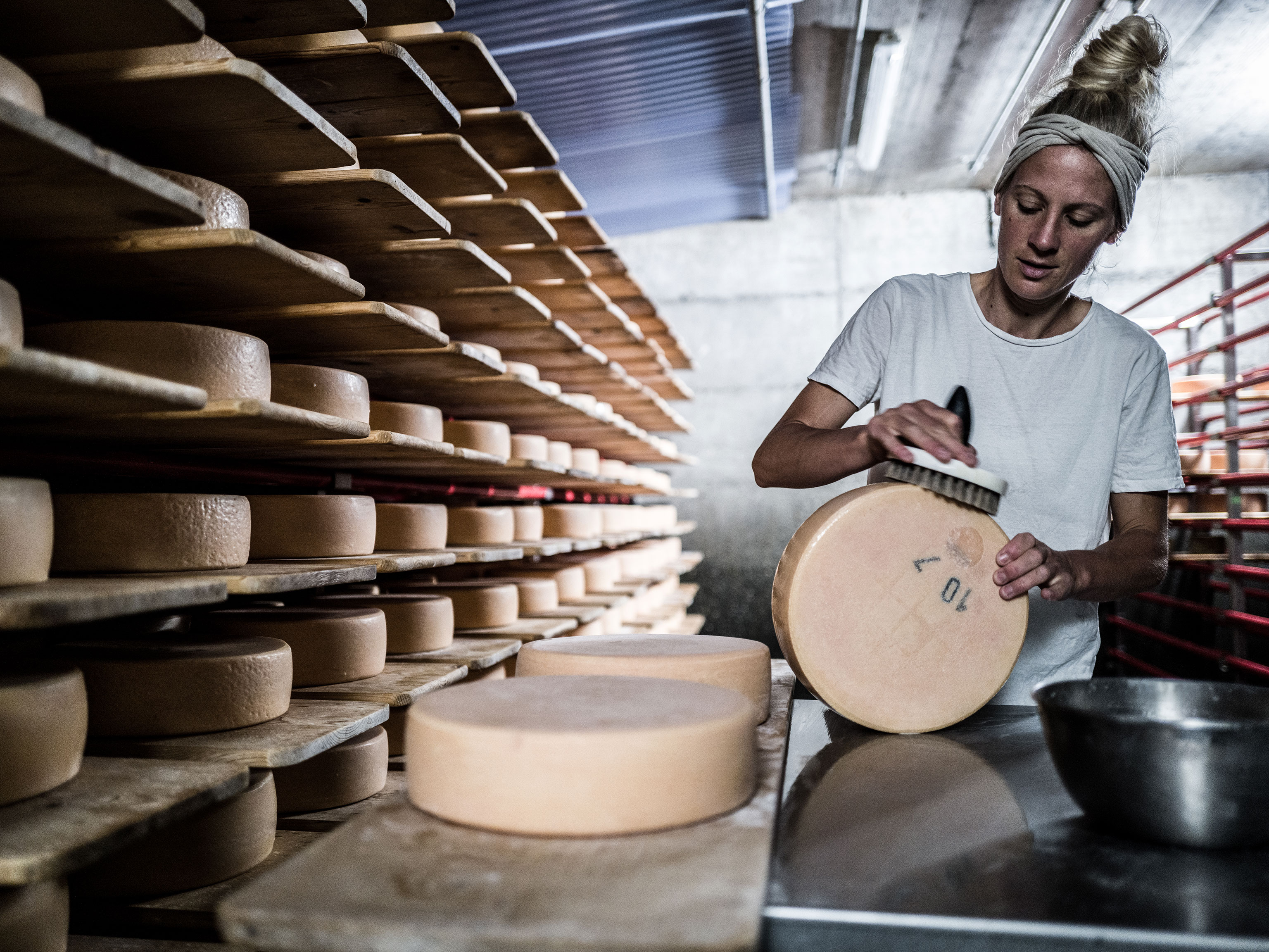 Katharina uses a large brush to brush a cheese wheel rind